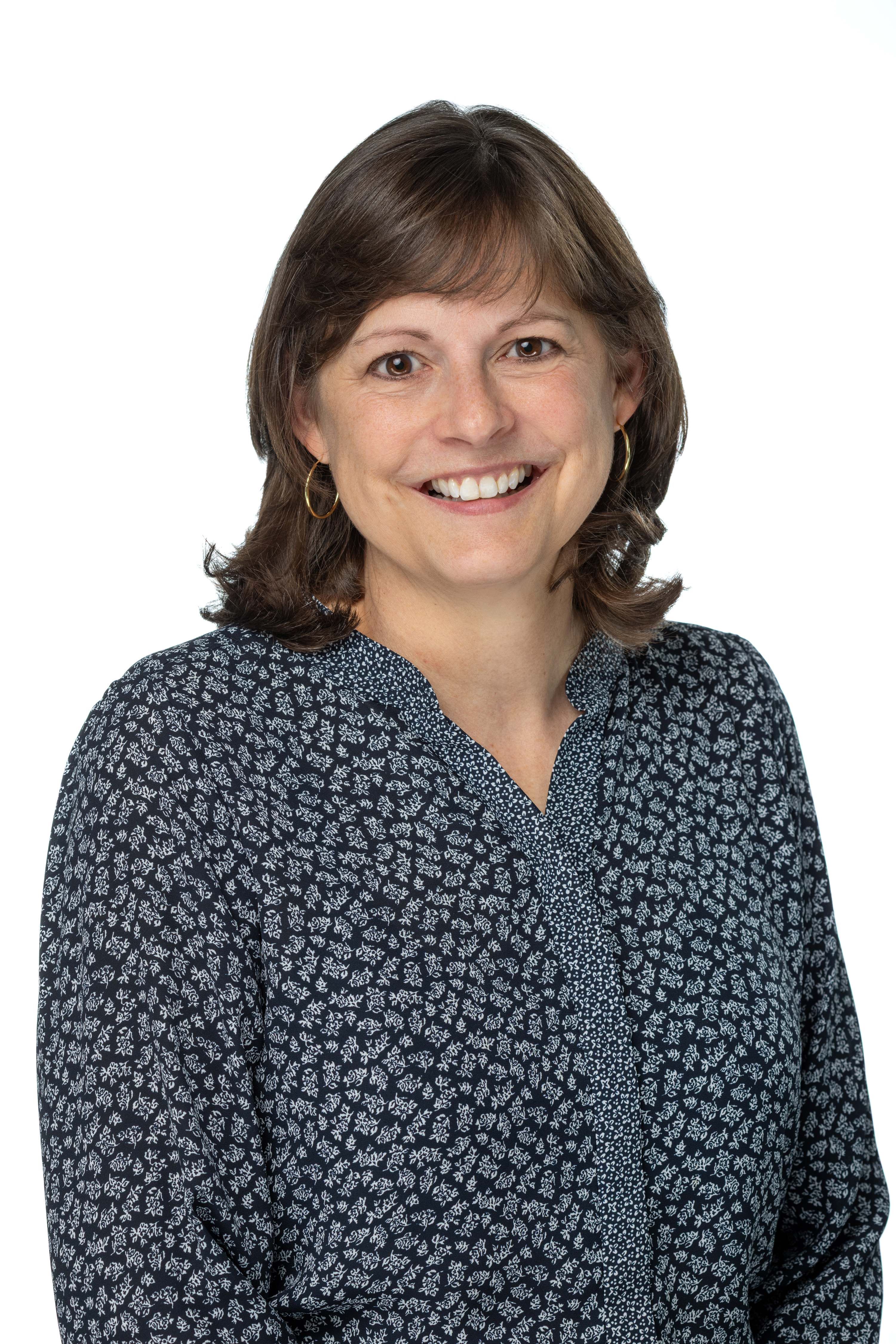 Maria V. Wathen, PhD, smiling and wearing a black top with a textured black-and-white jacket, gold hoop earrings, and a delicate gold necklace, against a white background.